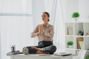 corporate businesswoman meditating in lotus pose with namaste gesture at workplace with Buddha head and incense stick clipart