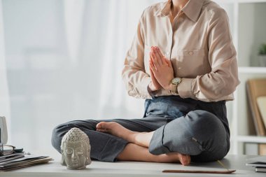 cropped view of businesswoman meditating in lotus pose with namaste gesture at workplace with Buddha head and incense stick clipart