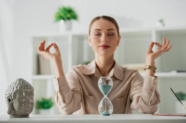 İş kadınının seçici odak noktası kapalı gözlerle meditasyon yapması ve iş yerinde Buda kafası, kum saati ve tütsü çubuğu ile Gyan Mudra.