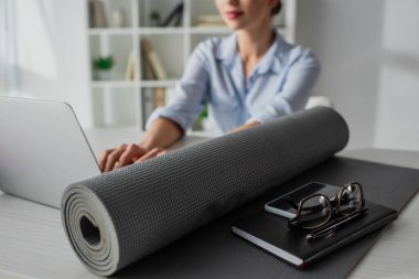 Yoga minderi, not defteri, akıllı telefon ve gözlüklü genç iş kadınının iş yerinde dizüstü bilgisayarda çalışmasının seçici odağı