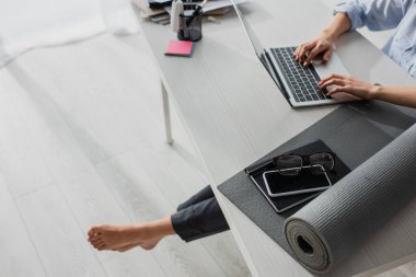 İş yerinde yoga minderi, not defteri, akıllı telefon ve gözlükle dizüstü bilgisayarda çalışan çıplak ayaklı iş kadını görüntüsü.