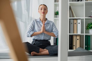 selective focus of businesswoman practicing yoga in lotus position with namaste gesture on mat in office  clipart