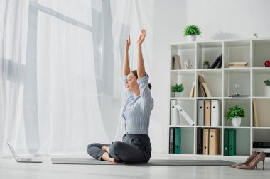 Genç iş kadınının dizüstü bilgisayar dersleri alması ve ellerinin havada Lotus pozisyonunda yoga çalışması. 