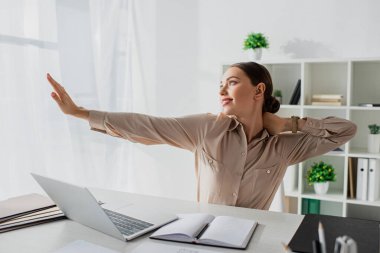 İş yerinde dizüstü bilgisayar ve not defteriyle kollarını açan genç ve güzel bir iş kadını.