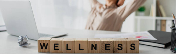 panoramic shot of businesswoman resting at workplace with laptop and alphabet cubes with wellness word 
