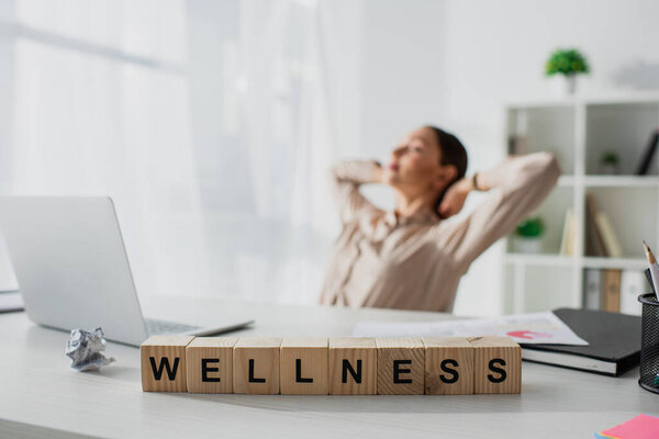 selective focus of businesswoman relaxing at workplace with laptop and alphabet cubes with wellness word 