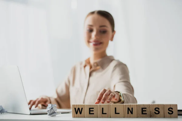 selective focus of happy businesswoman working with laptop at workplace with alphabet cubes with wellness word