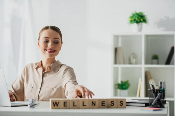 Smiling Businesswoman Sitting Workplace Laptop Alphabet Cubes Wellness Word — Stock Photo, Image