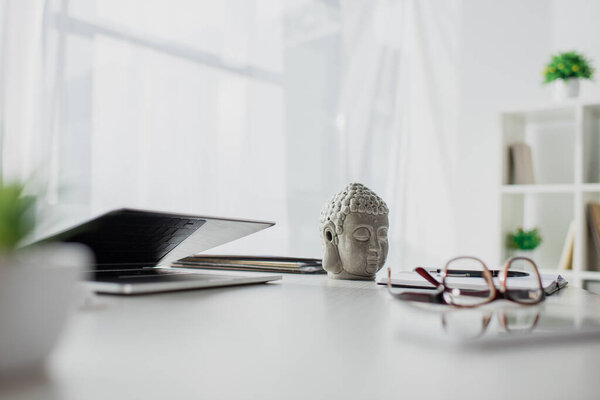 Buddha head, eyeglasses and laptop on table in modern office