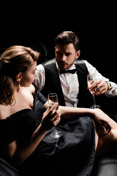elegant man and woman holding champagne glasses and looking at each other while sitting isolated on black