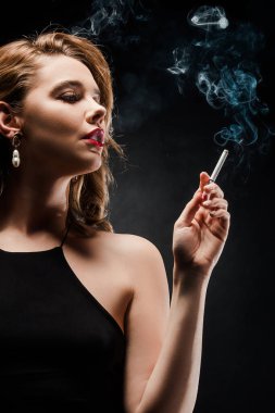 seductive, elegant woman looking away while holding cigarette on black background clipart