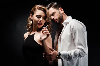 Beyaz düğmeleri olmayan yakışıklı adam siyah arka planda sigara tutan seksi, zarif kadına dokunuyor.