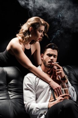 Şehvetli bir kız, elinde şampanya bardağı olan seksi bir adama sarılıyor ve siyah bir kanepede sigara içiyor.