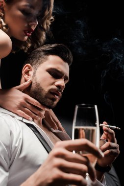 Yakışıklı, kendine güvenen bir adamın boynuna dokunan seksi kız elinde şampanya bardağı ve siyah bir sigara tutuyor.