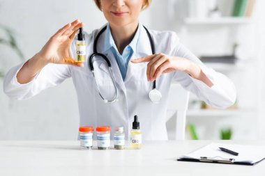 cropped view of doctor in white coat pointing with finger at bottle with cbd lettering  clipart
