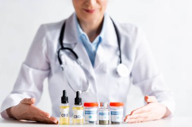 Doktorun ellerinde tıbbi esrar şişeleri ve CBD harfleri olan şişelere işaret ettiği görüntüler. 