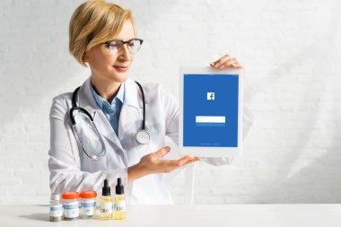 KYIV, UKRAINE - 5 Mart 2020: CBD ve tıbbi kenevir harfli şişelerin yanında facebook uygulaması ile dijital tablet tutan beyaz önlüklü olgun doktor