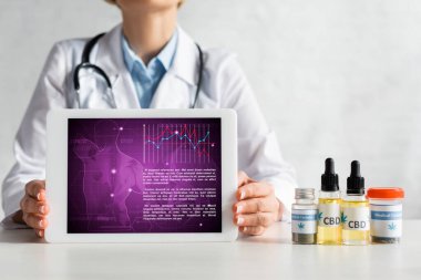 cropped view of mature doctor holding digital tablet with medical app near bottles with cbd lettering  clipart