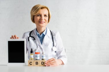Olgun doktor elinde dijital tablet ile CBD harfleri ve uyuşturucuları olan küplerin yanında boş ekran tutuyor 