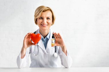 Neşeli ve olgun bir doktor elinde bir şişe CBD ile klinikte kırmızı kalp tutuyor. 