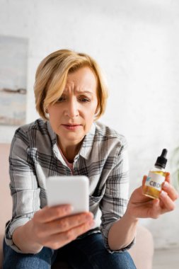 Seçici odak noktası olgun bir kadının elinde bir şişe dolusu cbd harfi ve akıllı telefon kullanması. 