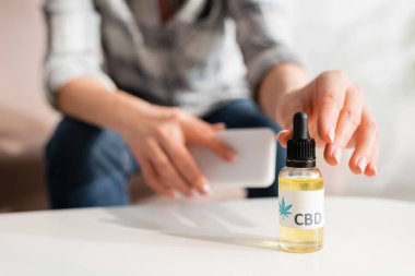 cropped view of mature woman taking bottle with cbd lettering and holding smartphone at home clipart
