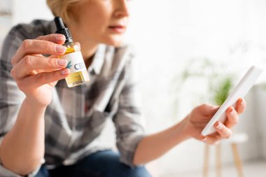 Olgun bir kadının elinde bir şişe cbd ile evde akıllı telefon kullanması görüntüsü.