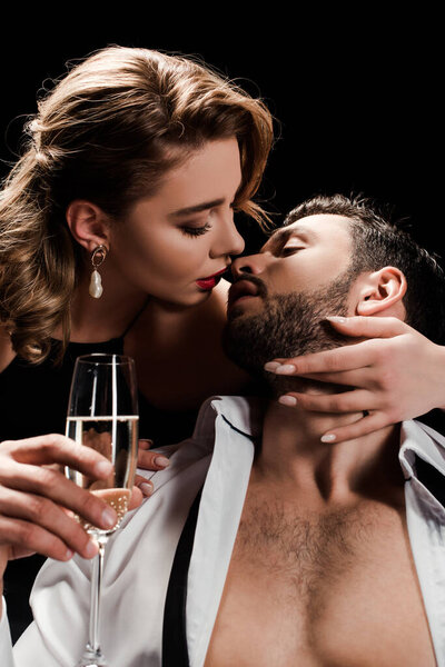attractive, elegant woman holding champagne glass and kissing handsome man isolated on black
