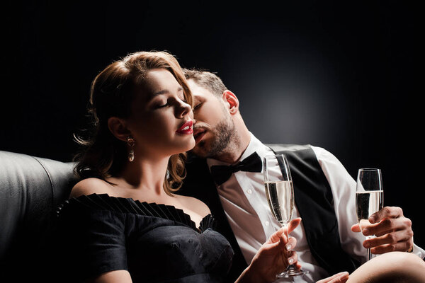 elegant man kissing seductive, beautiful woman sitting with closed eyes on black background