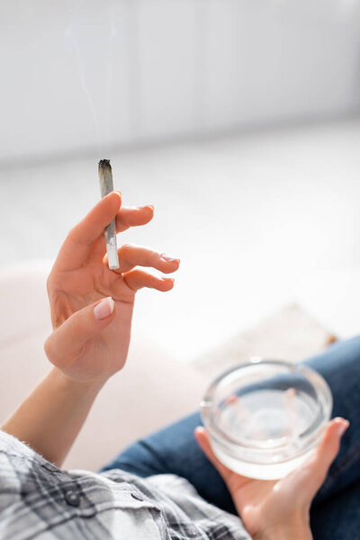 cropped view of mature woman holding joint with legal marijuana and ashtray 