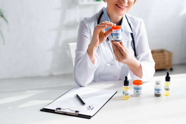cropped view of happy and mature doctor in white coat holding bottle with medical cannabis lettering 