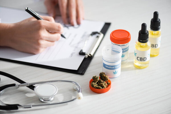 cropped view of doctor writing prescription near dried weed, bottles with cbd and medical cannabis lettering 