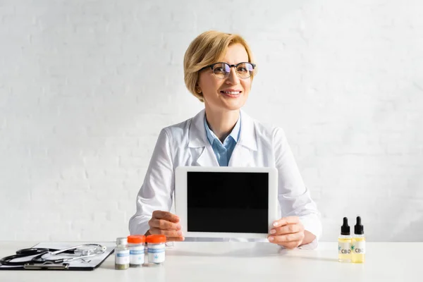 快乐的中年医生 在诊所的药瓶和医用大麻字母旁边 拿着空白屏幕的数码平板电脑 — 图库照片