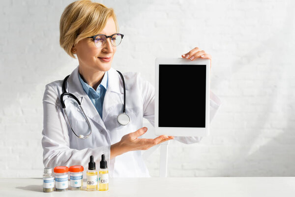 Врач среднего возраста в белом халате держит планшет с чистым экраном рядом с бутылками с CBD и медицинским каннабиса надписи в клинике
 