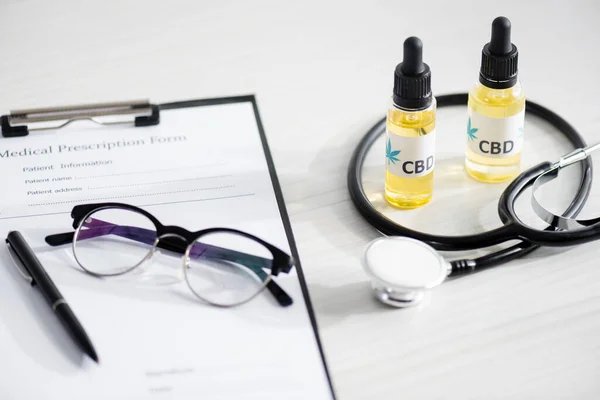 selective focus of bottles with cbd lettering near stethoscope, glasses and clipboard with medical prescription form