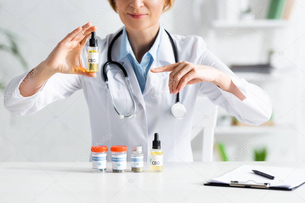 cropped view of doctor in white coat pointing with finger at bottle with cbd lettering 
