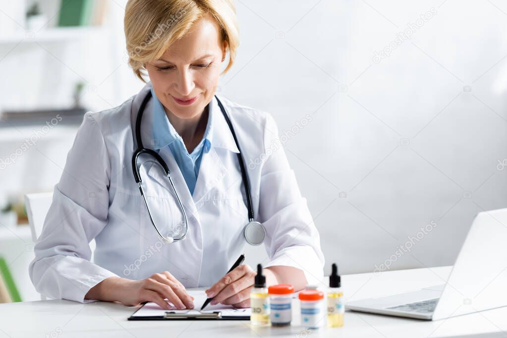 selective focus of cheerful and mature doctor writing prescription near bottles with medical cannabis and cbd 