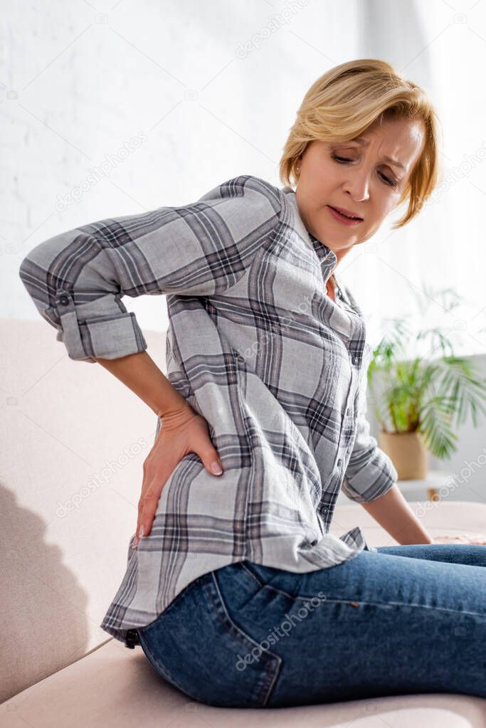 mature woman suffering from back pain and sitting on sofa 