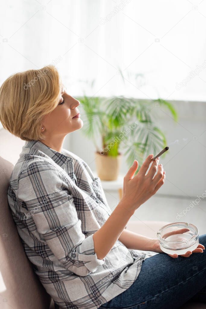 side view of mature woman smoking joint with legal marijuana and holding ashtray 