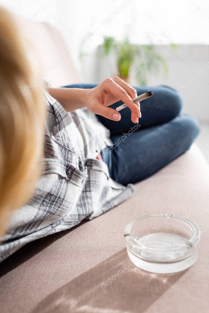 selective focus of mature woman lying on sofa and holding joint with legal marijuana near ashtray 