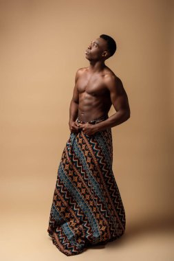 Battaniyeye sarılmış, bej üzerine poz veren seksi, çıplak afro adam.
