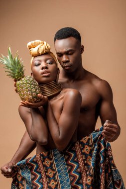 sexy naked tribal afro woman covered in blanket posing with pineapple near man on beige clipart