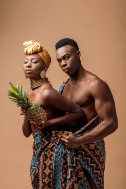Battaniyeye sarılı seksi çıplak afro kadın bej üzerine izole edilmiş bir adamın yanında ananasla poz veriyor.