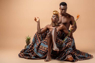 sexy naked tribal afro woman covered in blanket posing near man and pineapple on beige clipart