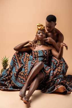 Battaniyeye sarılmış, erkeğin yanında poz veren ve bej üzerine ananas koyan seksi, çıplak afro kadın.