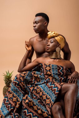 Battaniyeye sarılmış, erkeğin yanında poz veren seksi, çıplak afro kadın ve bej üzerine izole edilmiş ananas.