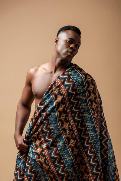 Sexy naked tribal afro man covered in blanket posing isolated on beige