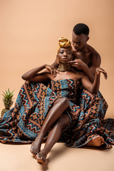 Sexy naked tribal afro woman covered in blanket posing near man and pineapple on beige
