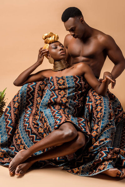 Sexy naked tribal afro woman covered in blanket posing near man on beige