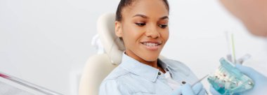 panoramic shot of happy african american woman in braces near dentist holding teeth model  clipart